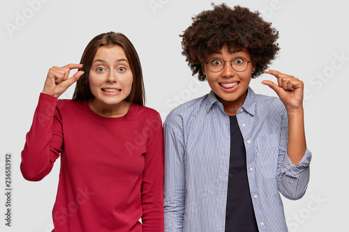 Fotografía Indoor shot of multiethnic young women gesture with hands, show something very little and invisible, demonstrate size gesture, dressed in fashionable clothing, isolated over white background