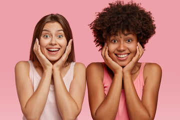 Wall Mural - Glad beautiful multiethnic girls hold chins, recieve good news from friend, have broad smiles, model over pink studio wall, express pleasant emotions and feelings. Interracial relationship concept