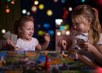 Wall Mural - Board game concept.Two little girls spend fun time at home playing board games. Board game field, many figures, two kid girls play in board game at home on dark blurred background