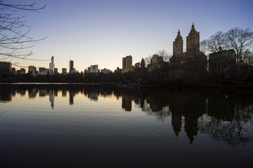 Wall Mural - Tranquil sunset view of the scenic skyline silhouette of the Upper West Side reflecting on the flat surface of the Central Park Lake on a calm winter evening in New York City