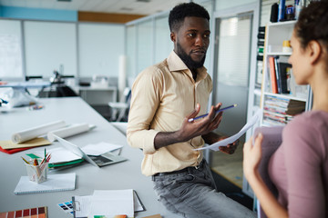 Young confident designer sitting on desk and explaining his view to colleague at meeting