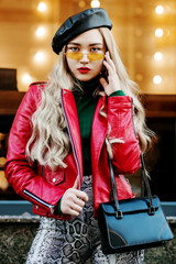 Wall Mural - Outdoor fashion portrait of young fashionable lady wearing red biker jacket, stylish yellow sunglasses, leather beret, trendy snake skin print trousers, holding small bag, posing in street of city