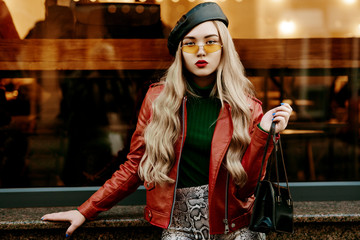 Wall Mural - Outdoor fashion portrait of young woman wearing red biker jacket, trendy orange sunglasses, leather beret, snake skin print trousers, holding bag, posing in street of city. Copy, empty space for text