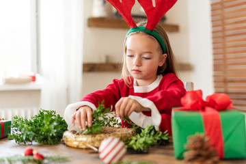 Cute preschooler girl dressed in reindeer costume wearing reindeer antlers making christmas wreath in living room. Christmas decoration family fun concept.