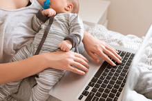 Busy Young Woman Working Or Study On Laptop Computer While Holding Her Baby In Arms At Home