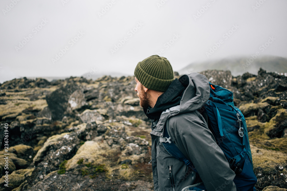 Young man in hiking trekking gear, waterproof jacket, green knit beanie and hike backpack walk through moss covered rough iceland terrain. Explore travel real wilderness lifestyle - obrazy, fototapety, plakaty 