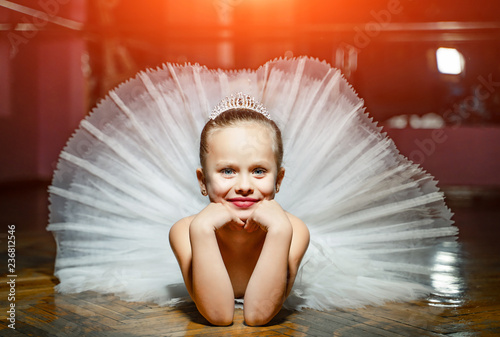 Fotografie, Obraz A portrait of cute smiling ballerina in white tutu and crown laying on the wooden floor with face in hands