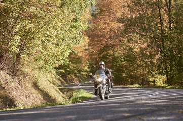 Bearded motorcyclist in helmet, sunglasses and black leather clothing riding cruiser bike along empty asphalt road winding among tall green trees on bright sunny summer day. Active lifestyle concept.