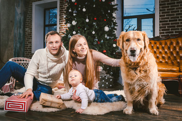 Wall Mural - theme Christmas and New Year family circle and domestic pet. Mom dad and child 1 year old Caucasian woman sitting on floor near Christmas tree with gifts and big dog breed labrador golden retriever