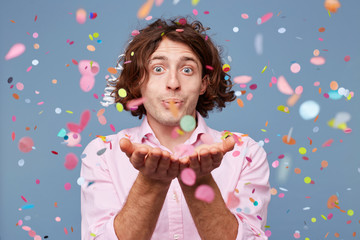 Close up of happy male blowing confetti off his hands. Party man gladly standing around falling down confetti, over blue background