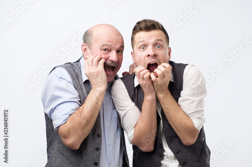 Fotografie, Obraz Emotive stupefied men stare at camera with bugged eyes full of fear, keep hands near opened mouthes