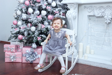 a little beautiful girl two years old dressed in a gray dress with a pink bow is sitting in a chair near an elegant New Year tree and straightening ruffles on a dress