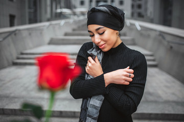 Wall Mural - Cheerful young arabian woman poses. She stands on stairs and look down. There is red rose in front of camera. It is unfocused. Model hold hands crossed.