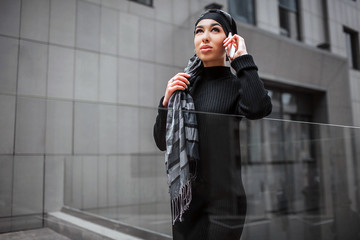 Wall Mural - Serious and concentrated arabian woman stand and pose. She talks on phone and look up. Model touch edge of hijab.