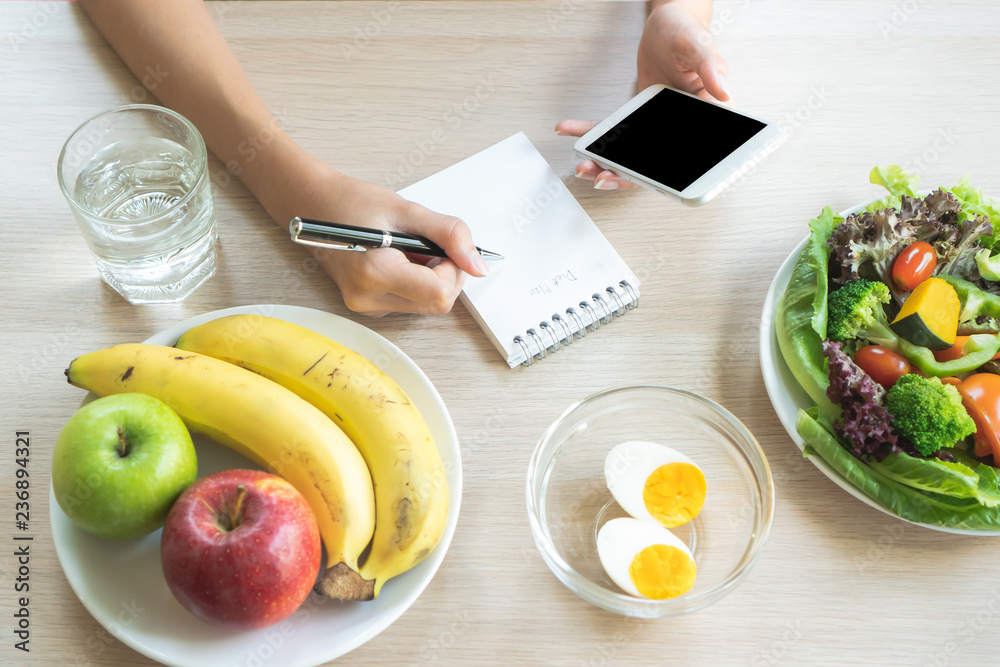 Dieting and calories control for wellness. Woman using smartphone calculate calories of food in breakfast during dieting for lose weight program and take notes. - obrazy, fototapety, plakaty 