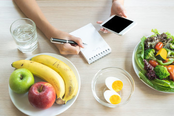 Dieting and calories control for wellness. Woman using smartphone calculate calories of food in breakfast during dieting for lose weight program and take notes.