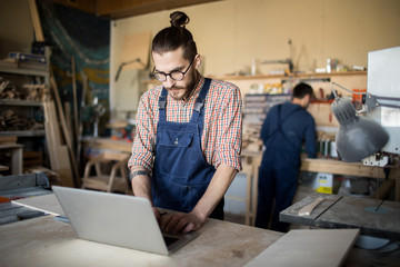 Portrait of workers in carpenters workshop, focus on modern man using laptop in foreground, copy space