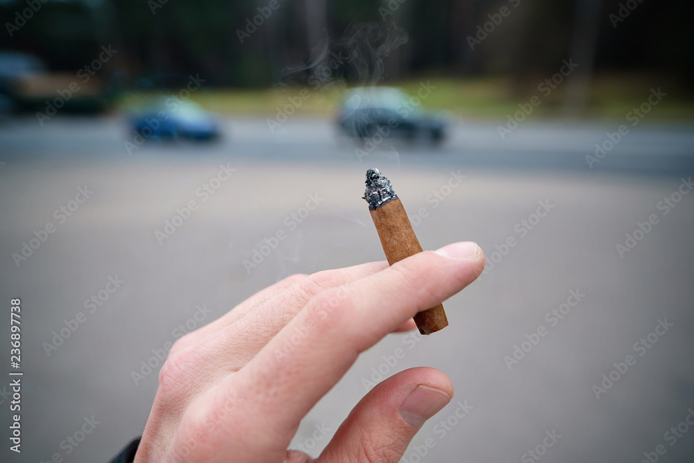 Photo Stock Man holding cigarette between fingers outside, on background of  road. Man holding cigarrillo in hand. Unhealthy lifestyle concept.  Unhealthy habit. selective focus. Nicotine addiction