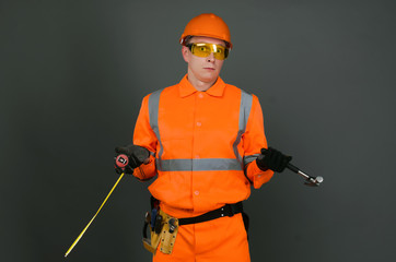 Guilty builder worker with a meter and hammer in hands justifies about lateness of construction and is shugging his shoulders isolated on gray background. Construction mistake.