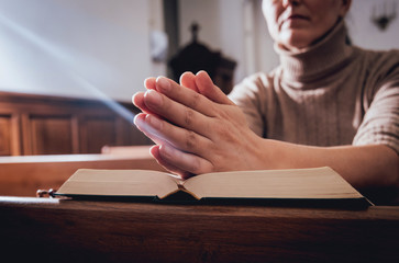 Wall Mural - Christian woman praying in church. Hands crossed and Holy Bible on wooden desk.