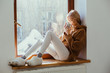 Portrait of blissful blond woman warming, relaxing sittng on window sill with hot coffee or tea in empty cozy home in sunny winter day.