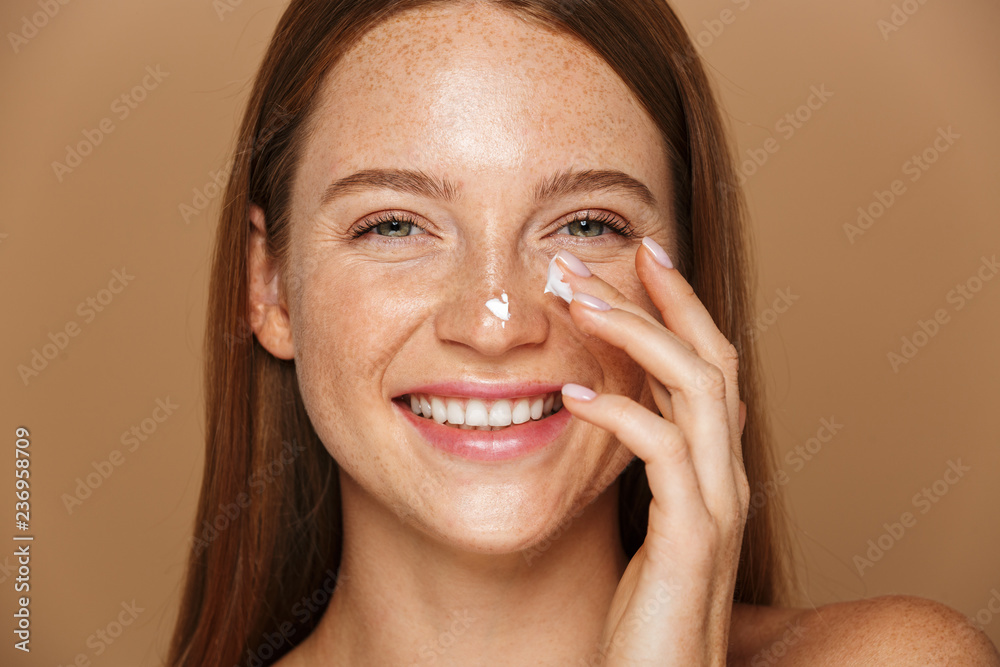 Beauty image of pretty shirtless woman smiling and applying face cream, isolated over beige background - obrazy, fototapety, plakaty 