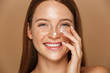 Beauty image of pretty shirtless woman smiling and applying face cream, isolated over beige background