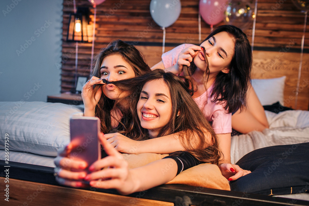 Three playful young women taking selfie on bed in room. Two models play ...