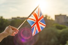 Hand Holding Great Britainin Flag An Open Window. Background Blue Sky, Silhouette Of The City, Sunset