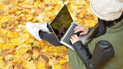 Wall Mural - Girl in hipster with laptop in autumn park. A woman in a cap using a laptop while sitting on fallen leaves. Freelancer in the hat uses remote communication technology. Remote work. View from above