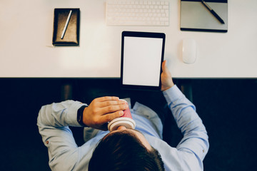 From above shot of unrecognizable male holding tablet with blank display and sipping hot drink while sitting at desk in office.Anonymous man with tablet drinking hot beverage in office