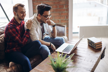 Two handsome young caucasian mates cooperate for making common project, using laptop, talk with each other. Business, team work and cooperation concept.