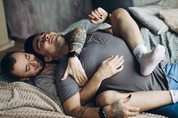 Wall Mural - Two homosexual male partners taking care of each other. Lovely gay couple have warm cuddle, having fun in bed and smile joyfully, feeling free and natural in same-sex conduct.