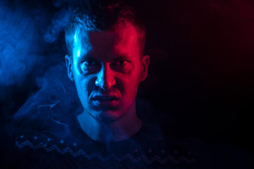 Wall Mural - Portrait of a man from close range who is angry and has bad emotions with a face mash and smoke around on a black isolated background. The face of the guy in the shade with color illumination.