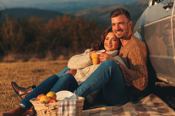 Wall Mural - 	 Beautiful young couple enjoying picnic time on the sunset. They drinking tea and sitting in a meadow leaning against a old fashioned car.