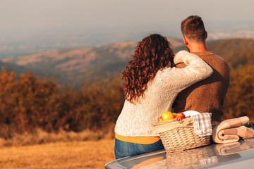 Wall Mural - Beautiful young couple enjoying picnic time on the sunset. They are sitting on old fashioned car and looking at distance.