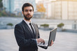 © speed300 - Businessman outdoors on a background of cityscapes holding a laptop looking straight.