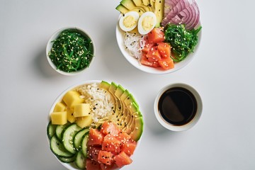 Raw Organic Poke Bowl with with rice, avocado, salmon, mango, cucumbers, chuka salad, quail eggs, sweet onions and soy sauce two plates close-up on white table background. Top view