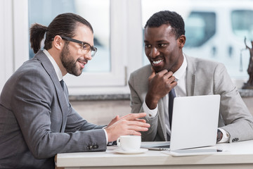 Sticker - smiling multicultural businessmen sitting at table with laptop and having discussion