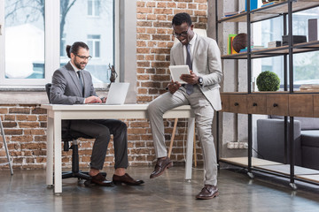 Wall Mural - handsome man typing on laptop keyboard and african american business partner sitting on table with digital tablet