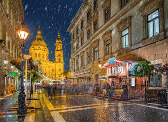 Budapest, Hungary - Snowy night at a Christmas market and shopping street with street-lamp, festive decoration and St.Stephen's basilica at background