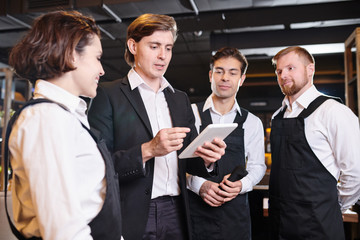 Briefing of waiters: serious confident handsome young manager using tablet and giving tasks to waiting staff while they preparing for banquet