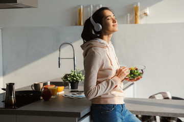 happy healthy woman in the kitchen standing daily morning routine eat salad listening music in headp