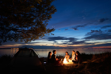 Night summer camping on sea shore. Group of young tourists sitting, laughing in high grass around bonfire near tent under beautiful blue evening sky. Tourism, friendship and beauty of nature concept.