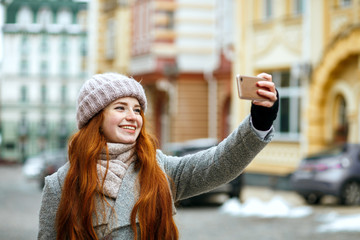 Wall Mural - Happy red head woman in winter apparel taking selfie on her mobile phone. Empty space