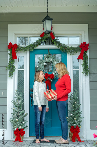 Fotografie, Obraz Mother and Daughter with a Christmas Gift Outside Their Home with a Festive Deco