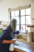 Mature Woman Cleaning Windows In Kitchen Using All Natural Cleaning Products