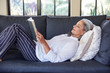 © Trinette Reed/Stocksy - Mature woman with grey hair reading a book lying down on sofa in living room