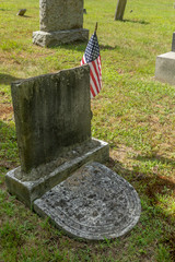 An old vertical grave stone - tombstone with obliterated writing stands in old cemetery with a small American flag on its right. Another curved grave stone sits at the base of the upright one.