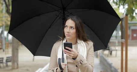 front view portrait of a frustrated woman checking weather app on a smart phone under the rain in wi